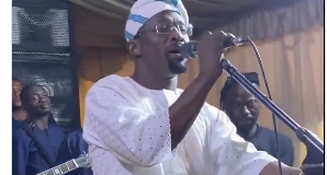 Malaika Warms Hearts After Sweet Gesture to Iya Rainbow at Event (Video) Male singer in white traditional outfit performs on stage with a live band behind; FASIWAJU TV logo overlay.