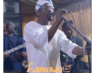 Malaika Warms Hearts After Sweet Gesture to Iya Rainbow at Event (Video) Male singer in white traditional outfit performs on stage with a live band behind; FASIWAJU TV logo overlay.