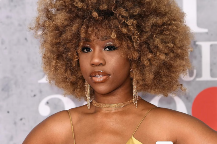 Portrait of a woman with a voluminous curly afro, wearing a gold spaghetti-strap dress and dangling earrings at a red carpet event.