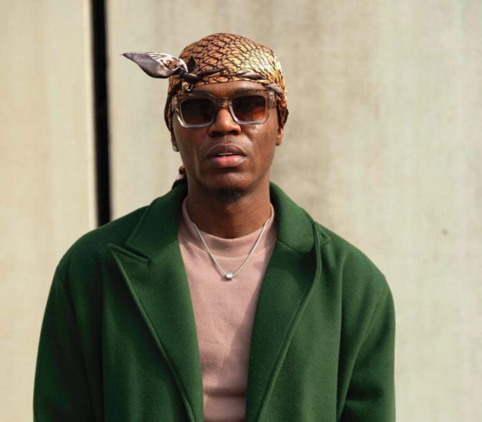 Spyro Man wearing a patterned headscarf and sunglasses, green overcoat, beige shirt, and a silver necklace, looking at the camera against a neutral background.