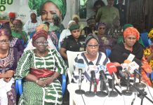 “We need our slots” – Iyaloja General demands political inclusion for traders Group of women in colorful traditional outfits seated at a press conference with microphones on a table in front of them; banners and onlookers in the background.