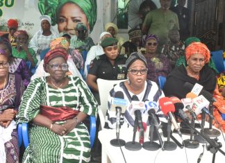 “We need our slots” – Iyaloja General demands political inclusion for traders Group of women in colorful traditional outfits seated at a press conference with microphones on a table in front of them; banners and onlookers in the background.
