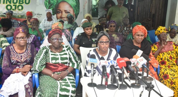 Group of women in colorful traditional outfits seated at a press conference with microphones on a table in front of them; banners and onlookers in the background.
