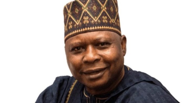 Smiling man in a patterned cap and dark traditional attire against a white background, portrait.