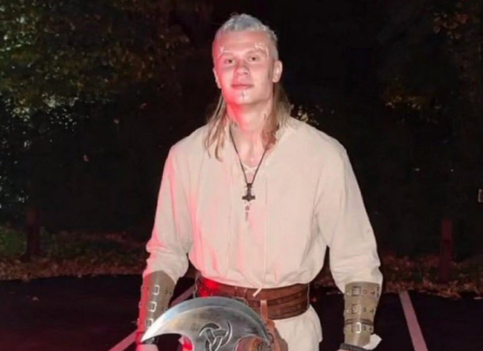 Person in medieval cosplay stands outdoors at night, wearing a beige tunic, leather bracers, belt, and holding a round shield with a cross emblem