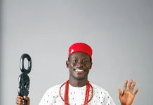 “You Be Black Jesus” — Geh Geh Blasts Blessing CEO Over Alleged Fundraising Drama Smiling man in a white traditional outfit with red cap and red bead necklaces, holding a ceremonial staff and raising his other hand in greeting.