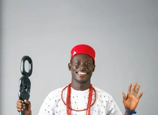 “You Be Black Jesus” — Geh Geh Blasts Blessing CEO Over Alleged Fundraising Drama Smiling man in a white traditional outfit with red cap and red bead necklaces, holding a ceremonial staff and raising his other hand in greeting.