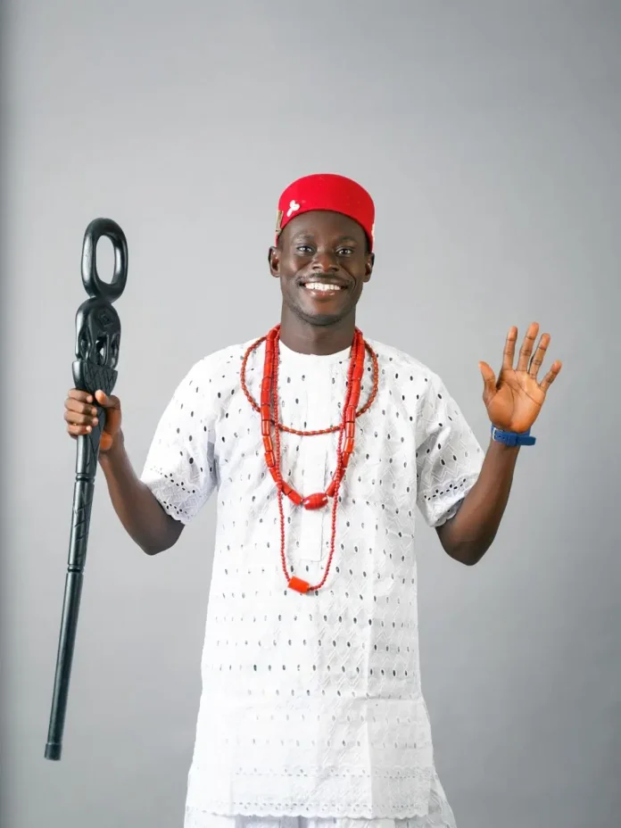 Smiling man in a white traditional outfit with red cap and red bead necklaces, holding a ceremonial staff and raising his other hand in greeting.