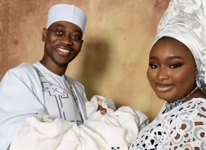 A joyful couple in traditional white attire cradling a newborn baby wrapped in white cloth.