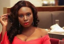 Mo Bimpe overwhelmed by tears of joy as she breaks silence on triplet miracle Woman in a red off-the-shoulder dress sits at a restaurant table, looking at the camera with red lipstick and long wavy hair reveals earrings in a dining setting.