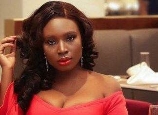 Mo Bimpe overwhelmed by tears of joy as she breaks silence on triplet miracle Woman in a red off-the-shoulder dress sits at a restaurant table, looking at the camera with red lipstick and long wavy hair reveals earrings in a dining setting.