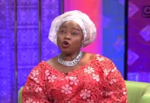 Yetunde Wumi Breaks Down Over Lack of Formal Education Woman wearing a white head wrap and red floral dress speaks on a TV talk show set with colorful backdrop.