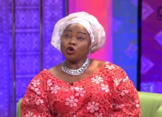 Yetunde Wumi Breaks Down Over Lack of Formal Education Woman wearing a white head wrap and red floral dress speaks on a TV talk show set with colorful backdrop.